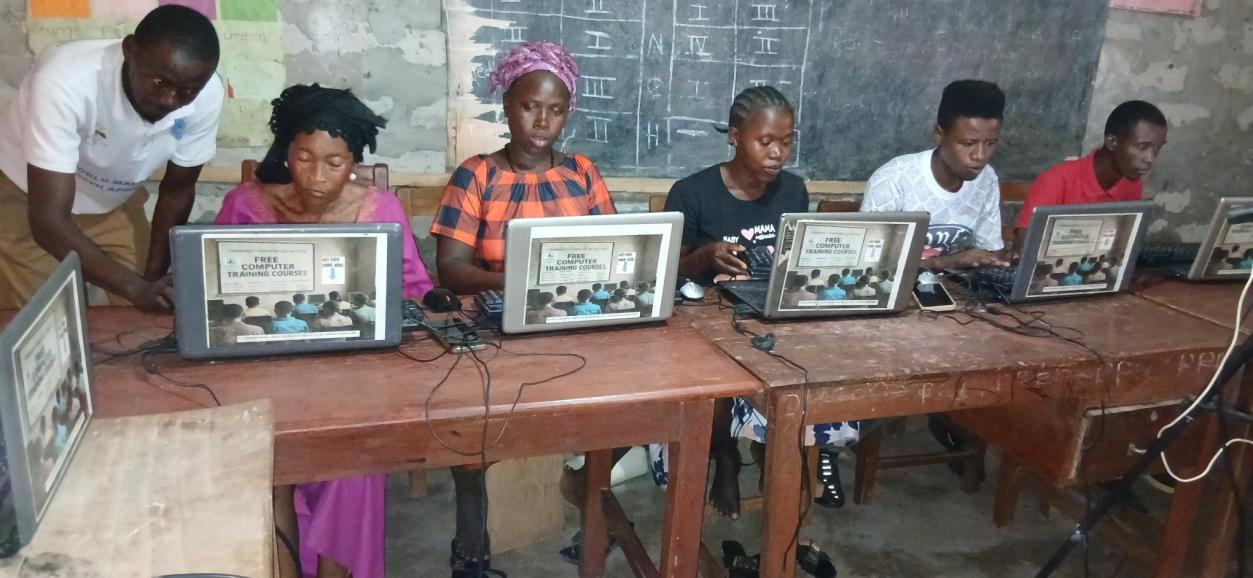 A facilitator supporting community members during free computer training in a rural classroom